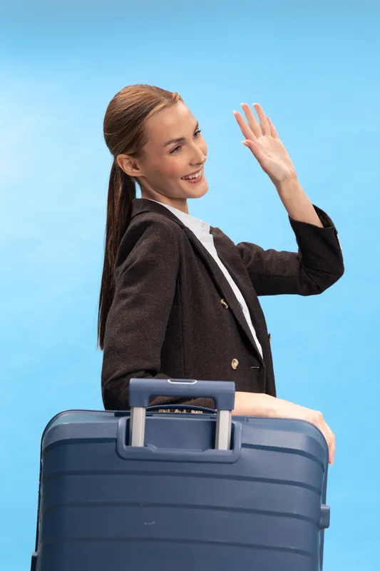 Young woman with long hair in black blazer smiles and waves while holding a blue suitcase handle against a light blue background. Concept for travel, vacation, and business trip.