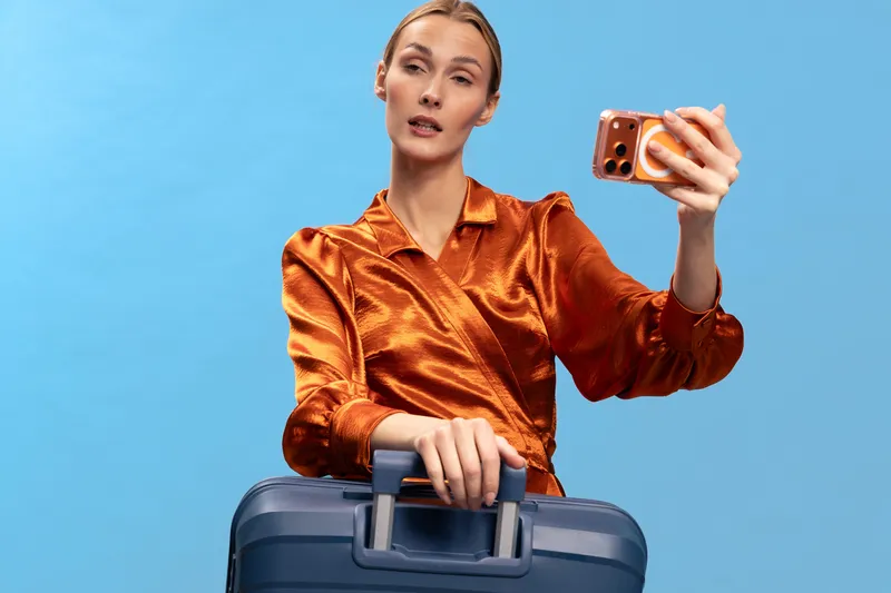 Young woman in shiny orange blouse takes selfie with smartphone while holding blue suitcase on blue background. Concept for travel, fashion, and modern lifestyle.
