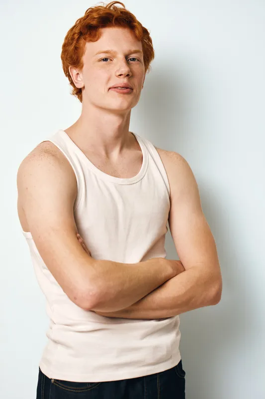 Young man with red hair and fair skin wearing sleeveless shirt standing with crossed arms against white background. Casual style and confident expression in studio portrait.