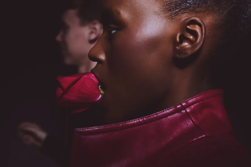 Profile of a young black woman with dark skin holding a red rose in her mouth wearing a red leather jacket. Concept of beauty, fashion, and floral elegance with a dark background.