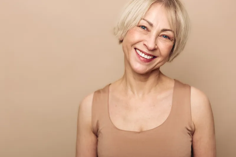 Smiling mature woman with short blonde hair wearing sleeveless beige top stands against neutral background. Concept of happiness, beauty, aging gracefully, confidence, and natural skin.