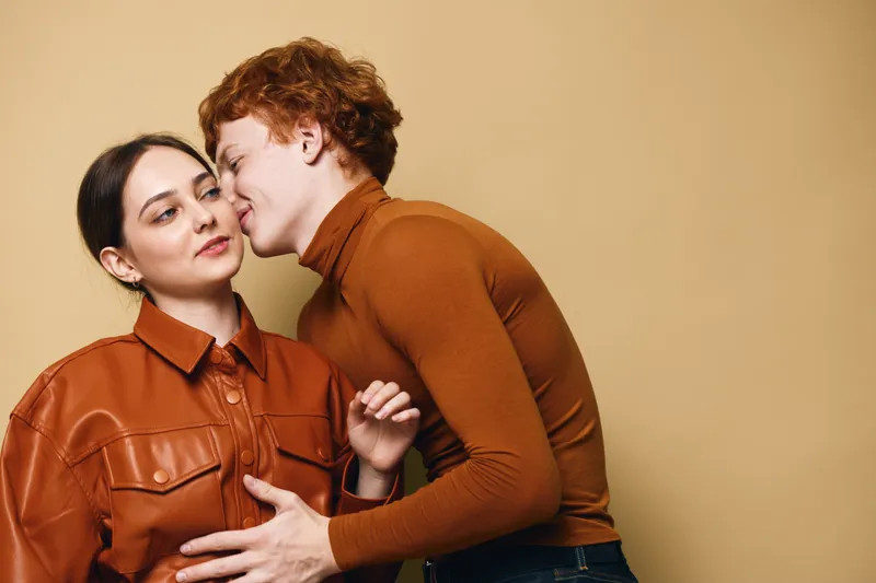 Young couple in casual clothing, man kissing woman on cheek with affection and warmth, romantic moment with love and connection indoors on beige background.