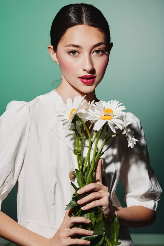 Fashion model in white blouse holds a bundle of daisies. Her makeup includes bold lips and blushed cheeks. Studio shot with soft contrast on teal background, beauty concept.