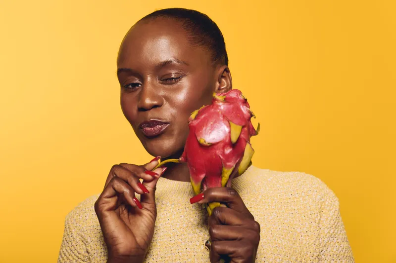 Closeup of a smiling black woman holding a dragon fruit near her face on yellow background. Healthy lifestyle concept with fresh exotic fruit and natural beauty of African ethnicity.