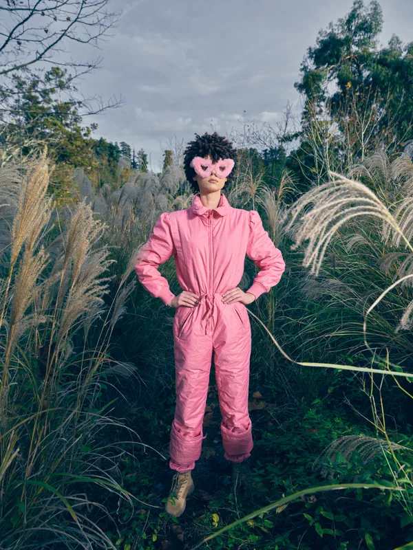 Pink jumpsuit woman outdoors in a field of tall grasses, showcasing playful fashion and quirky heart glasses. A bold whimsical portrait with vivid color and creative style in a natural meadow setting.