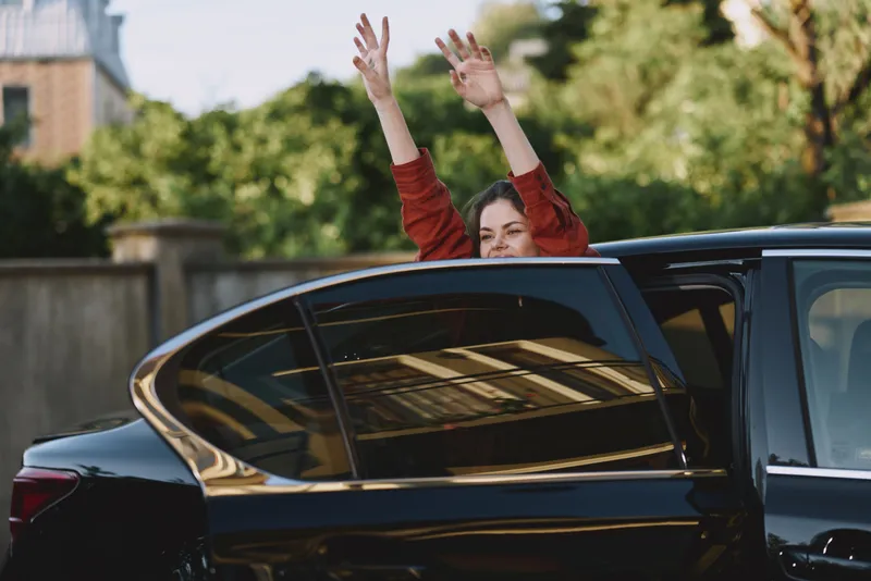Confident woman leans from a sleek car window, arms raised in joy. Bright daytime street scene captures carefree urban living, travel dreams, and personal freedom