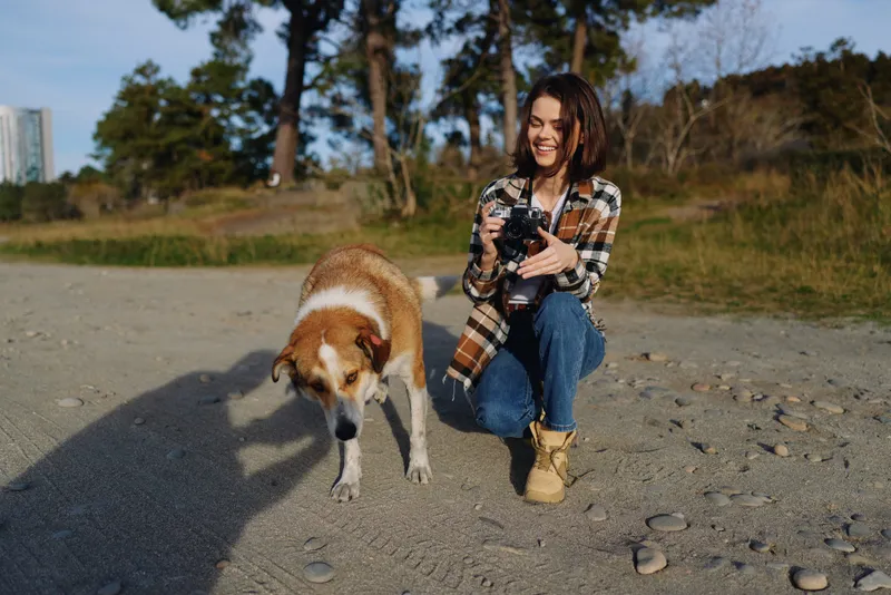 Woman photographer kneels on a sandy beach with her dog, holding a camera and smiling, capturing candid outdoor moments in a natural coastal setting.