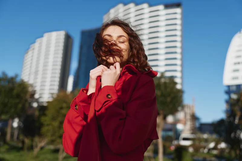 Portrait of a woman in a red coat outdoors, urban setting with tall buildings, wind in hair, cheerful mood, bright sky, candid street scene.