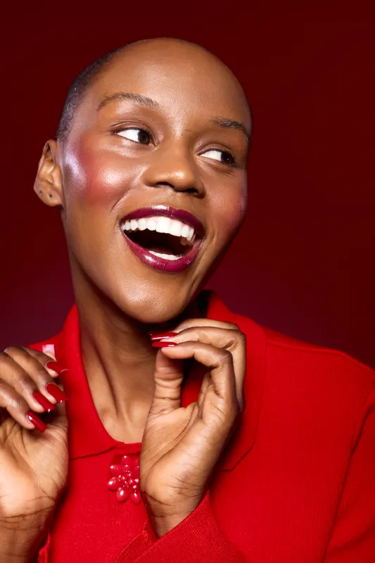 Happy African woman with dark skin smiles broadly showing white teeth, wearing red clothes and nail polish, posing on red background. Concept of joy, beauty, fashion, and confidence.