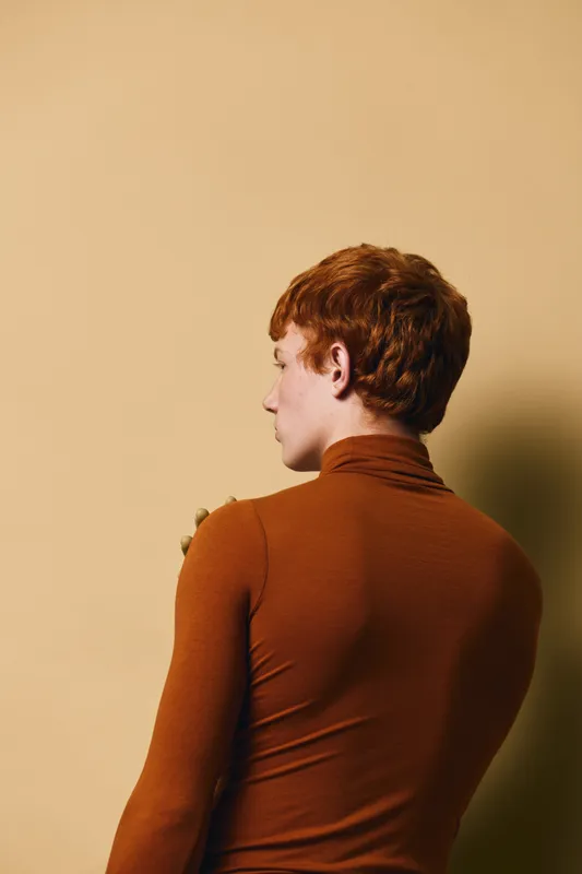 Young person with red hair wearing brown turtleneck stands against beige background looking sideways with profile visible and soft shadow on wall.