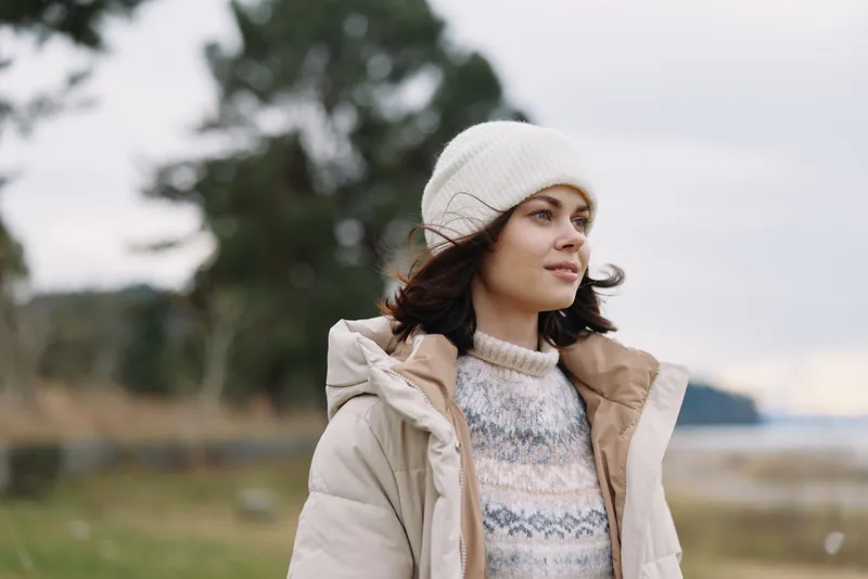 Woman in a warm beige coat and knit sweater standing outdoors with a beanie, calm landscape behind, capturing a chilly day mood and subtle sense of adventure.