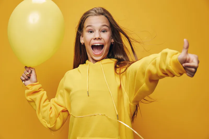 Girl in yellow hoodie holding balloon with excited expression and thumb up gesture. Happy teenager celebrating with festive atmosphere and joyful mood on bright background.