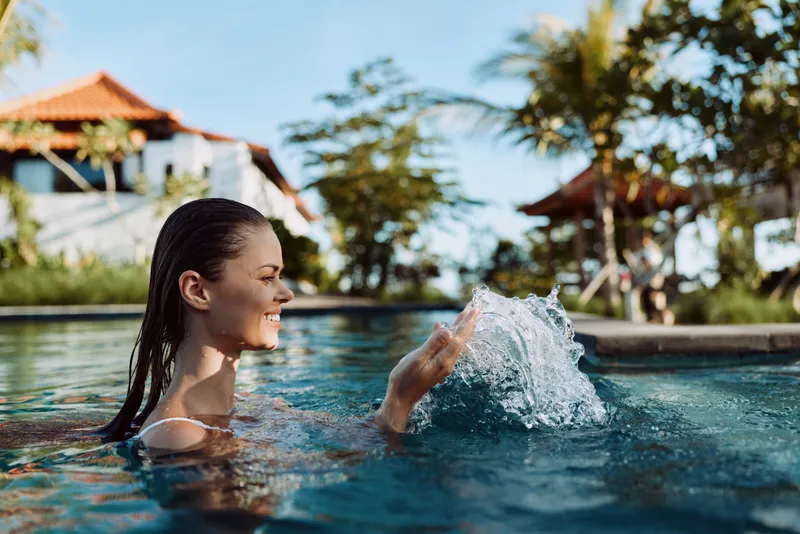 Cheerful woman in an outdoor pool, smiling as water splashes, warm sunlight and tropical backdrop creating a relaxed leisure moment with a vacation vibe and bright summer energy
