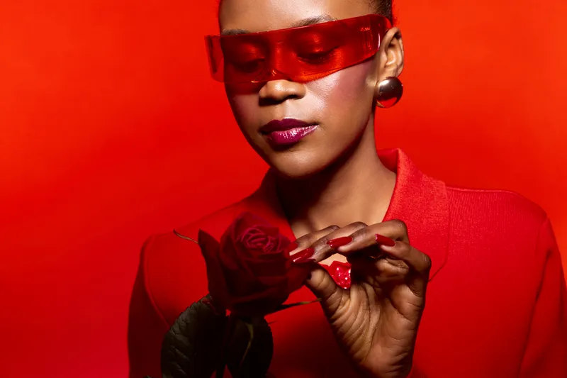 Stylish African American woman with dark skin wearing red sunglasses and red outfit holds red rose against vibrant red background. Concept of fashion, beauty and color matching.
