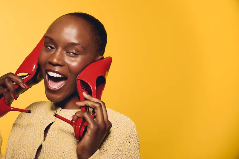 Happy African woman with dark skin holds red high heels near face, smiling joyfully on yellow background. Concept for fashion, beauty, footwear, and positive emotions.