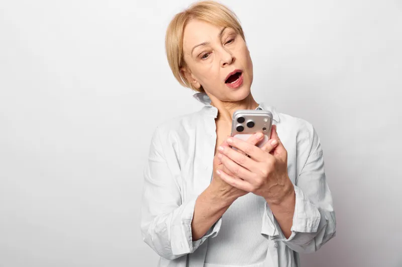 Blonde woman holding a smartphone with surprised expression in a clean studio portrait, communication and news concept, soft light, neutral white background today