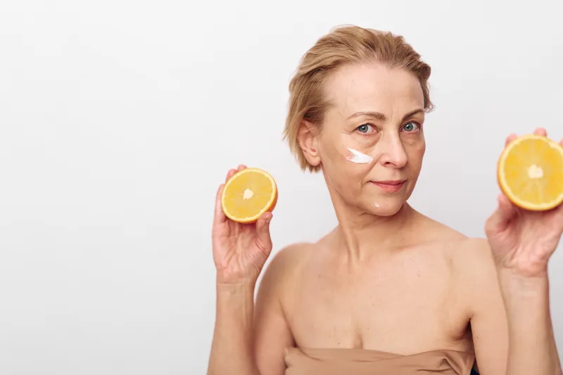 Mature woman with short hair applies natural cream on face holding fresh orange halves in both hands on white background. Concept of skincare, antiaging, and organic beauty treatment.