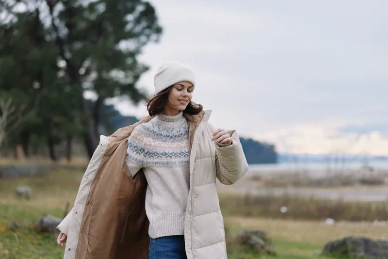 Woman outdoor winter coat jacket beanie clothing fashion moment in a chilly landscape as she adjusts her coat and sweater, enjoying a calm breeze and natural scenery