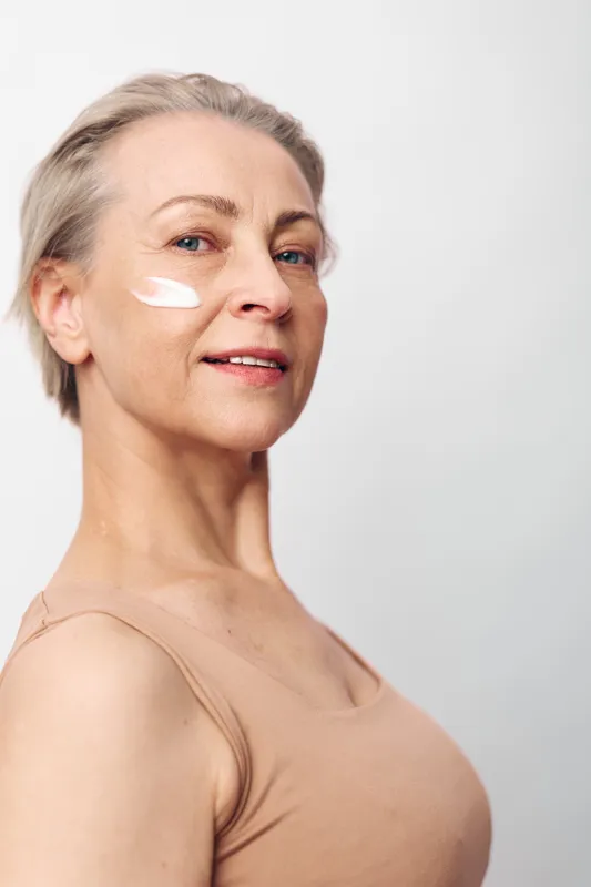 Mature woman with short hair applies white cream on face, smiling gently. Skincare, beauty, and self-care concept with natural skin and minimal makeup on light background.