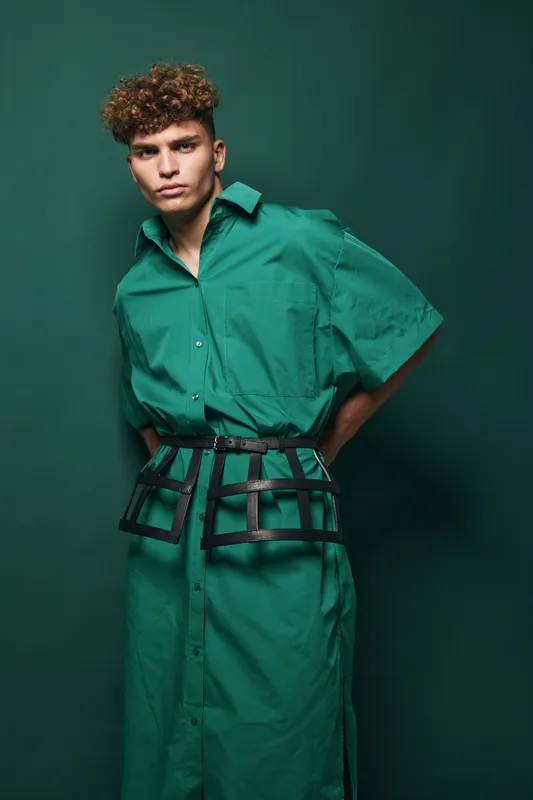 Young man with curly hair wearing green shirt dress and black decorative cage belt, posing for portrait with confident expression isolated on green background in studio.