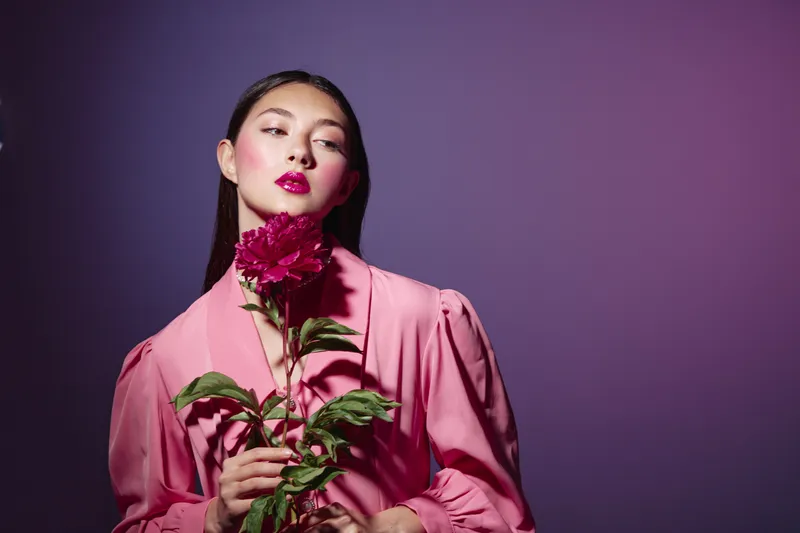Fashion model dressed in a pink blouse holds bright pink flowers, posing against purple studio background. Creative beauty and vibrant fashion concept with strong color contrast.
