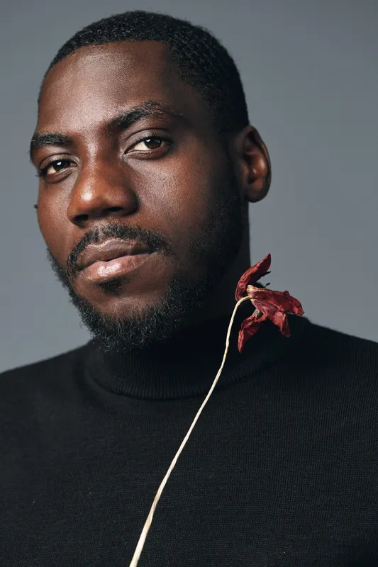 African man portrait with beard and short hair wearing black turtleneck holding dried red leaf in creative conceptual visual style with serious expression on gray background.