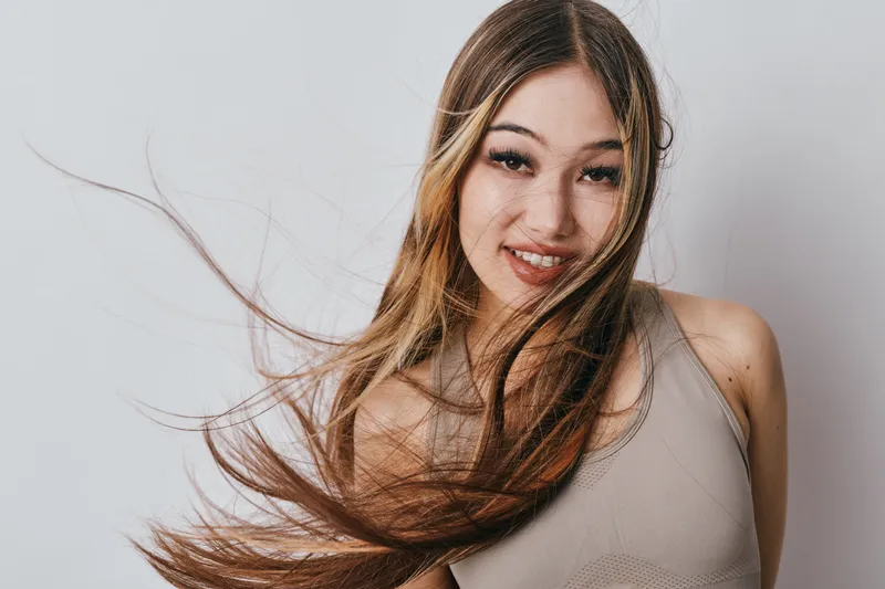 Young woman with long hair in motion wearing a beige top smiling with eyes looking at camera on a plain white background studio fashion portrait.