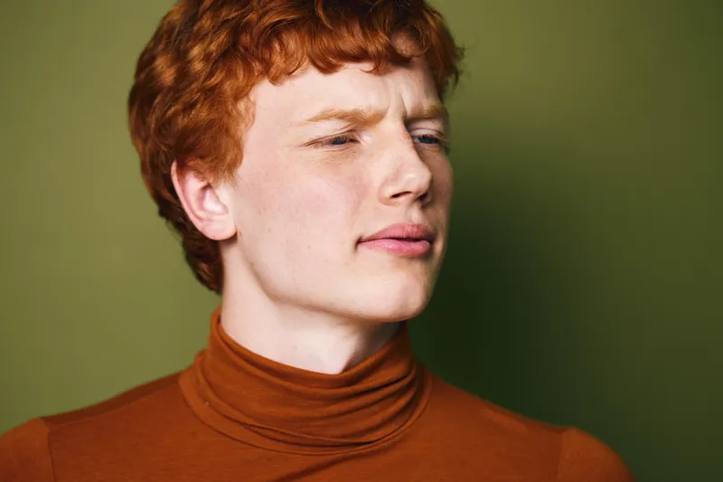 Young man with red hair wearing brown turtleneck sweater looking away on green background, casual style and thoughtful expression in studio portrait photography.