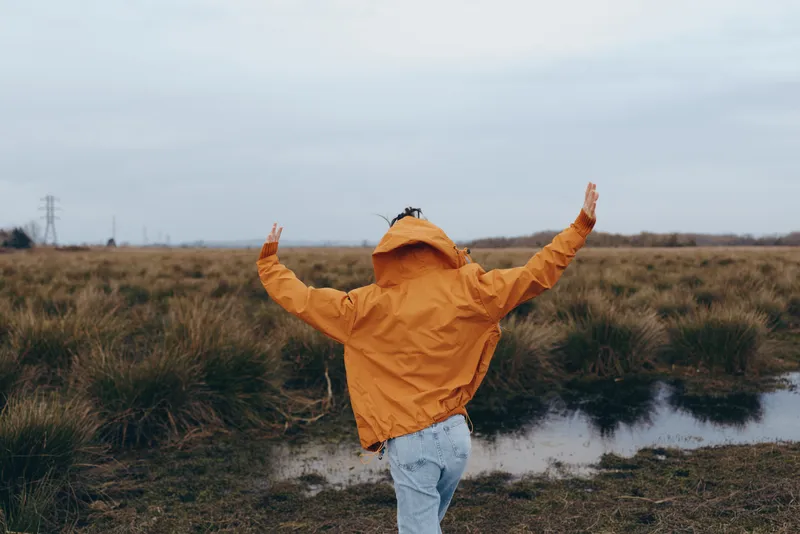 Woman lifestyle outdoors in a grassy field beside a calm river, orange hooded jacket, arms raised in joy, windy posture, cloudy daylight, natural colors, serene mood