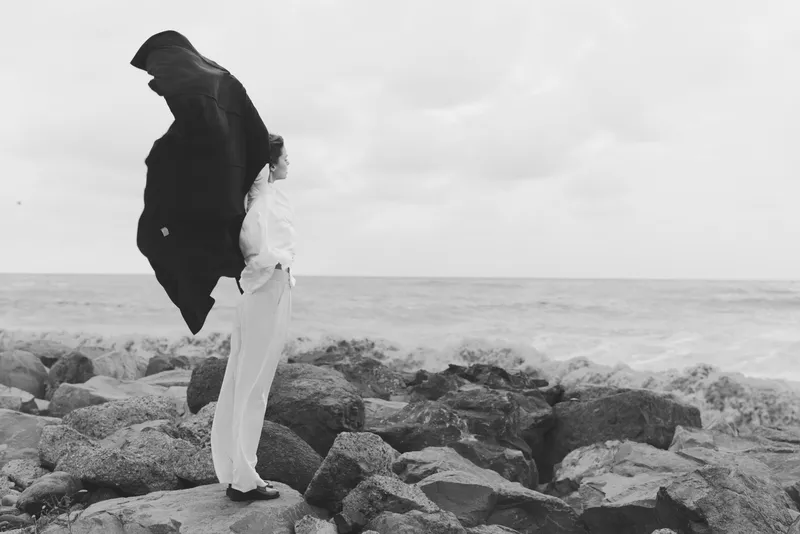Cloaked, hooded figure stands on a rocky coastal shore as waves crash, capturing mood, solitude, and wind by the sea