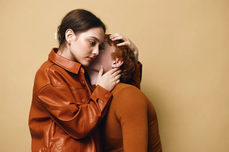Young woman comforting man with intimate embrace showing support and care, both wearing brown clothing against beige background in emotional connection and empathy.