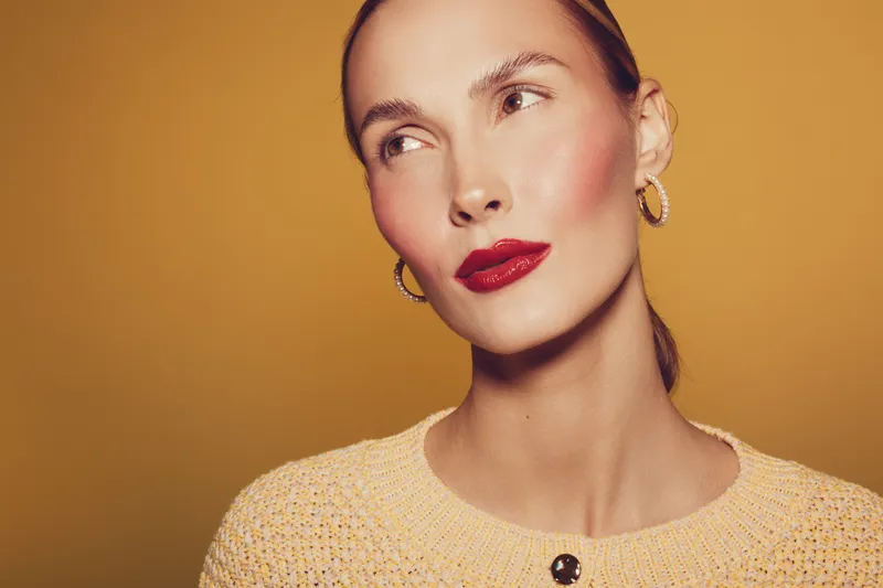 Portrait of a stylish woman with red lipstick and gold hoop earrings wearing a beige sweater. She looks away thoughtfully on a warm orange background. Fashion and beauty concept.
