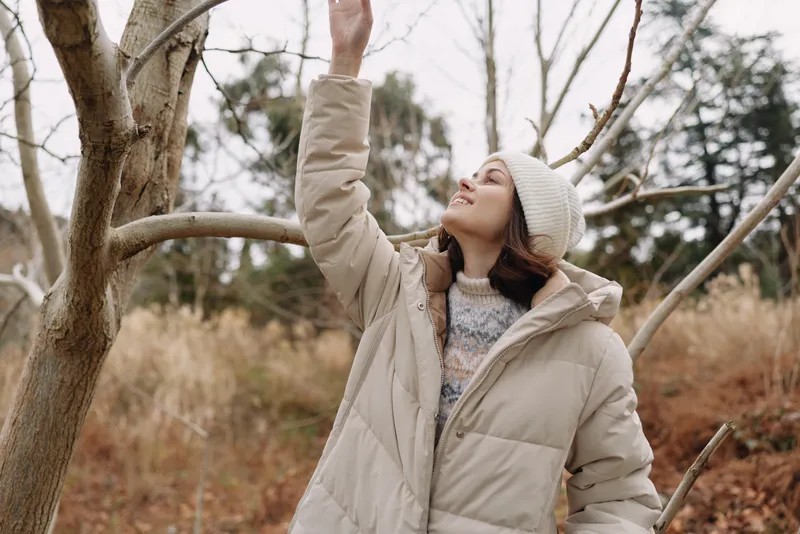 Woman in a beige puffer coat reaches toward a bare tree in a chilly autumn park, knit hat and soft light create a cozy outdoor moment highlighting winter fashion and nature.