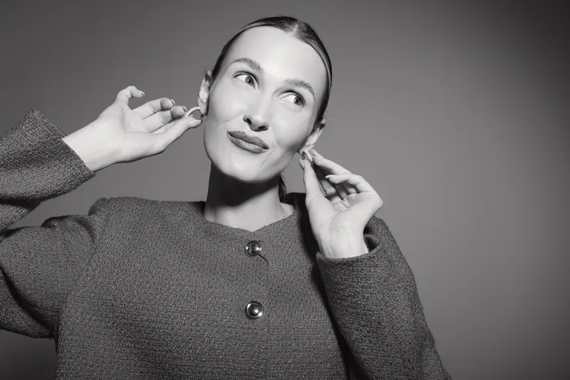 Young white woman with fair skin adjusts earrings, wearing textured coat in studio with neutral background. Concept for fashion, accessories, beauty, and elegance.
