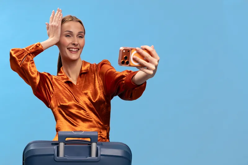 Young woman in orange blouse takes selfie with smartphone while standing behind suitcase on blue background. Concept of travel, vacation, and modern communication.
