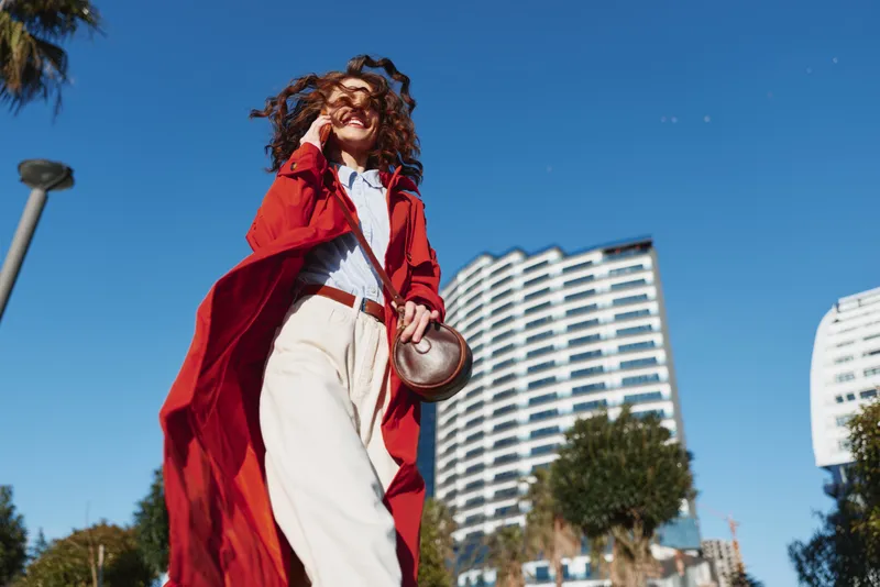 A young woman with curly brown hair strides outdoors in a bold red coat, white trousers, and a round bag against a sunny city backdrop with modern buildings behind her.
