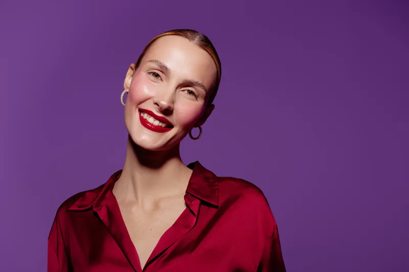 Smiling woman with red lipstick and hoop earrings wears a red satin blouse against purple background. Portrait of confident female model with natural makeup and bright studio lighting.