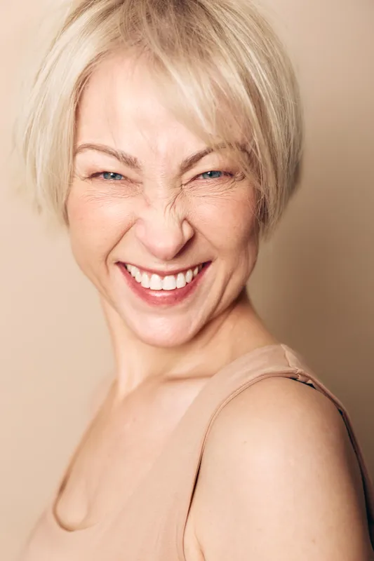 Blonde woman with short hair smiles widely showing teeth, eyes squinted, wearing beige top on soft beige background. Concept of joy, happiness, emotion, and natural beauty close-up.