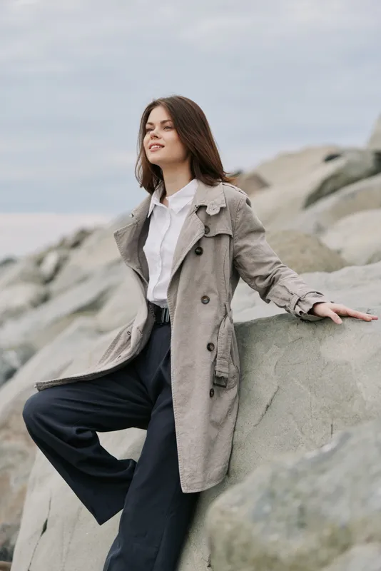 Young woman in a long coat sits on large rocks by the sea, looking relaxed and confident. The cloudy sky and natural stones create a calm outdoor mood and fashion concept.