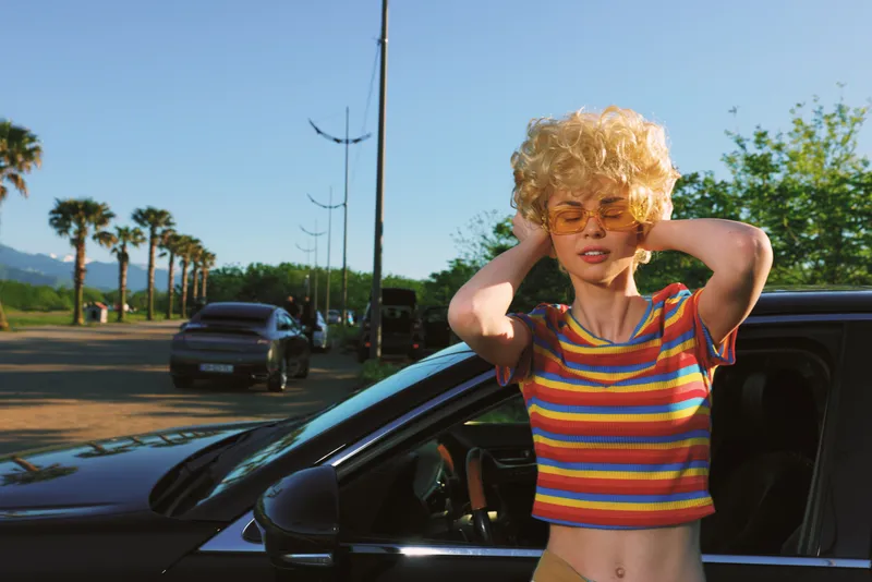 Woman with blonde curly hair wearing a striped rainbow crop top stands beside a parked car on a sunlit street, retro film color mood, casual lifestyle moment, confident pose