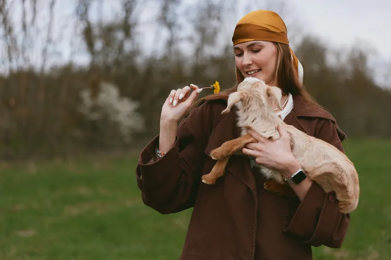 Lifestyle scene of a smiling woman outdoors with a goat kid, feeding it a leaf, warm brown jacket and orange bandana. A candid moment of bonding, care, and gentle affection in a grassy park.