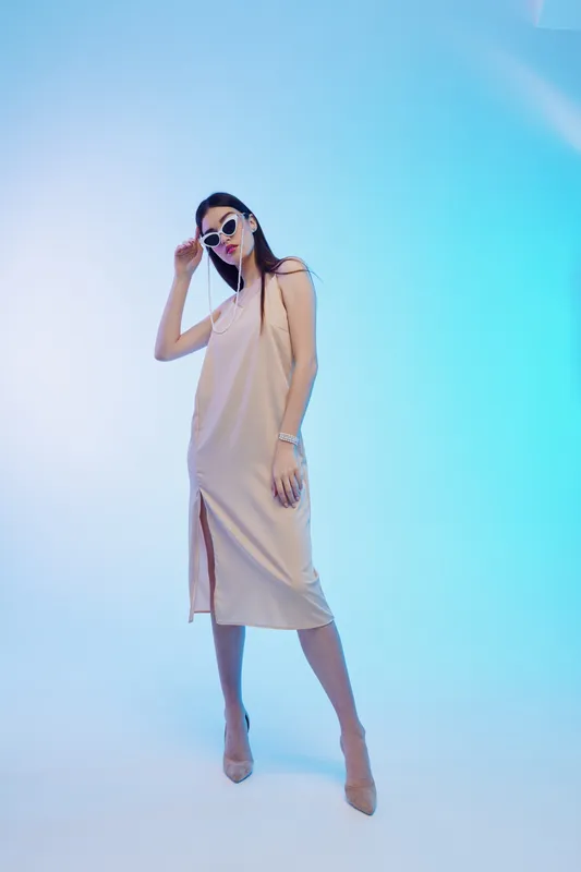 Stylish woman in a beige dress and sunglasses poses confidently against a blue gradient background. Concept of woman beauty fashion and modern elegance with soft lighting.