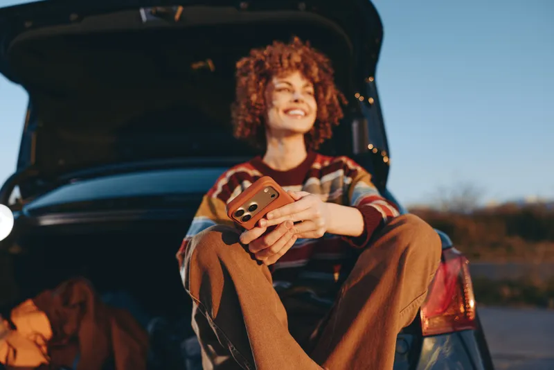 Cheerful woman lifestyle outdoors near a car at sunset, holding a smartphone with both hands, curly hair softly lit, smiling and relaxed, warm golden light, cinematic blur