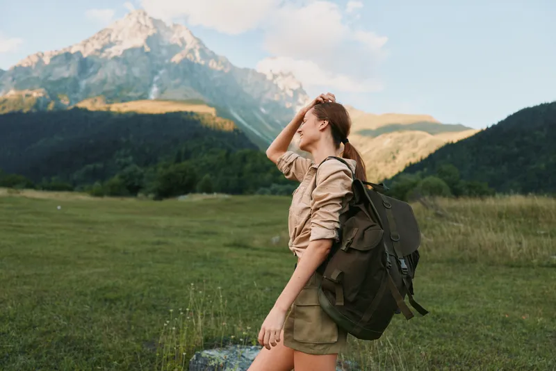 Young woman with backpack enjoys sunlit meadow as distant mountains rise, capturing a candid moment of adventure, freedom, and connection with nature.