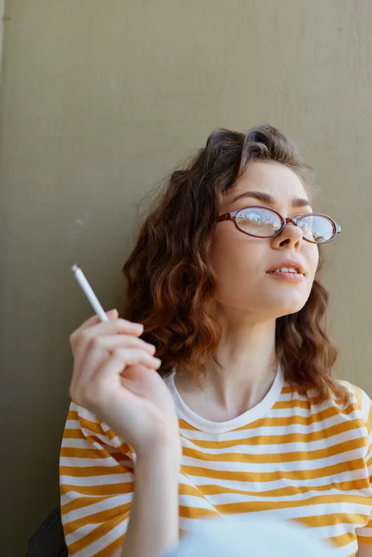 Casual woman wearing striped shirt and glasses, smoking gently, relaxed pose, soft light, home vibe, candid everyday moment captured in natural colors and calm mood