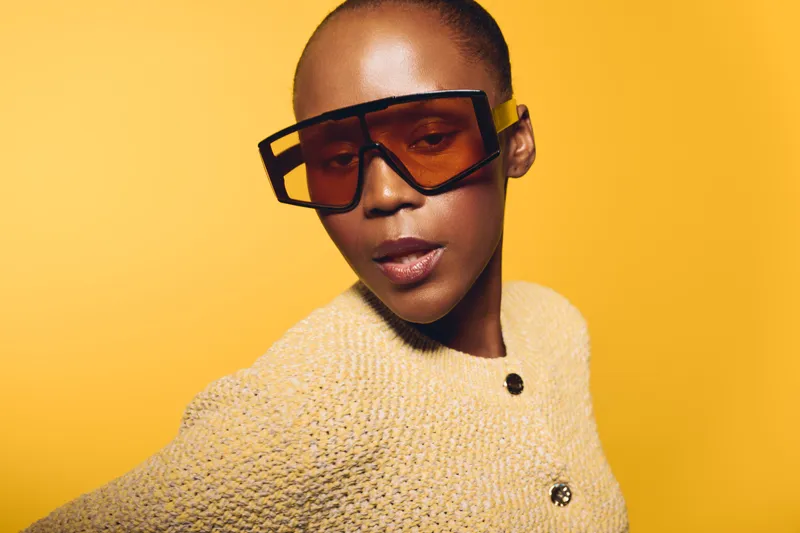 Elegant dark skinned African woman wearing oversized fashion sunglasses and a textured beige sweater poses confidently against a bright yellow background. Concept of style and beauty.
