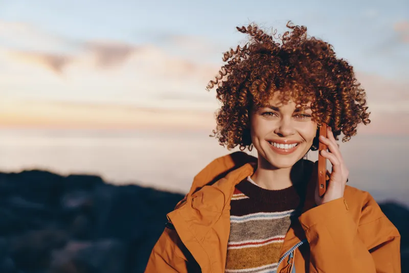 Woman lifestyle portrait outdoors at sunset, curly hair, smile as she holds phone to her ear, orange jacket and striped sweater, warm evening light
