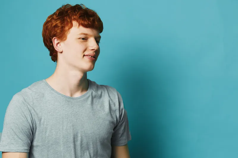 Young man with red hair wearing a gray t shirt standing against blue background looking to side with slight smile in casual studio portrait photography.