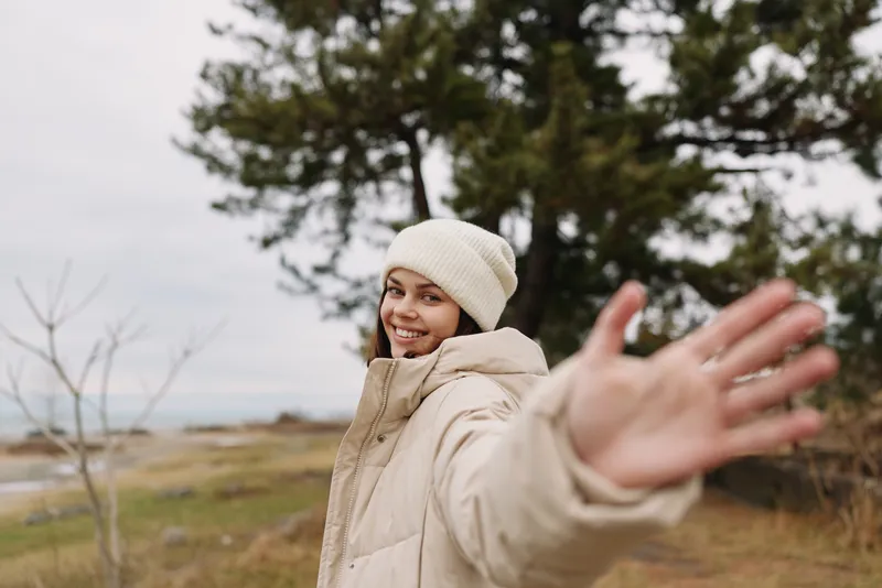 Outdoor portrait of a smiling woman in a beige jacket and beanie extending her hand toward the camera, with a scenic natural background and soft light, conveying warmth, friendliness, and carefree