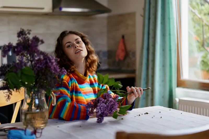 Smiling woman in a rainbow striped sweater arranges lilac flowers on a dining table by a sunlit window, creating a warm, cozy home mood, and a sense of calm, everyday creativity.